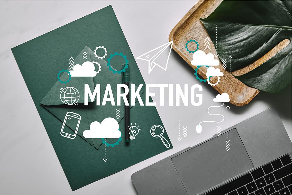 A flat lay of a work desk featuring green stationery, a clipboard, a laptop, and a wooden tray with a leaf, overlaid with various marketing icons and the word "MARKETING" in bold white letters.